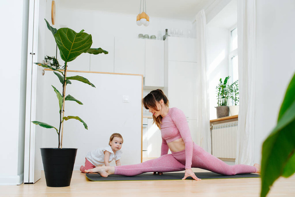 Young mother doing exercises at home dropping into the splits, next to crawls her little daughter - Valokuva, kuva