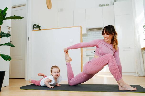Young flexible mother engaged in stretching workout with baby daughter at home in the background of the kitchen - Foto, Imagem