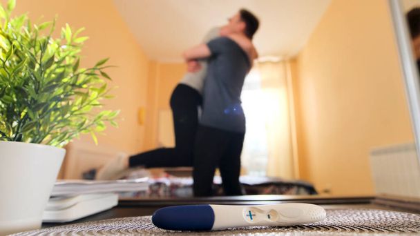 Close up of positive pregnancy test lying on a table and woman embracing a man. Stock footage. Happy husband hugging his pregnant wife in the room, family concept. - Photo, Image