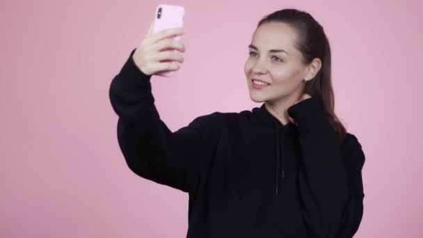 Woman smiles and makes selfie on mobile phone, isolated on pink background - Záběry, video