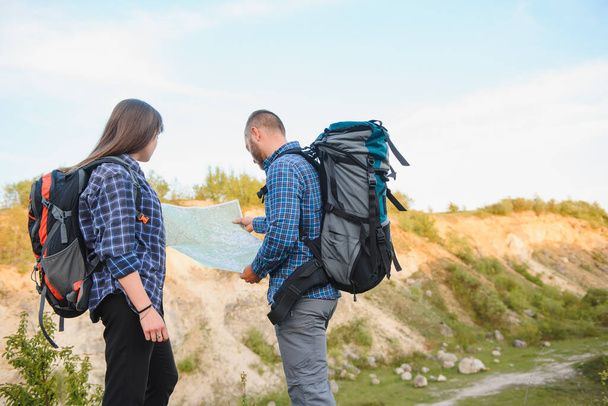 Beautiful couple of a travelers are searching way on location map while standing on high hill in sunny day, male and female hikers are walking together in mountains during long awaiting summer weekend - Photo, Image