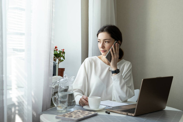 Businesswoman consults by phone and drink coffee at home. Talented manager remote working in laptop reading e-mail. Remote working and telephone consultations concept. View in window - Φωτογραφία, εικόνα