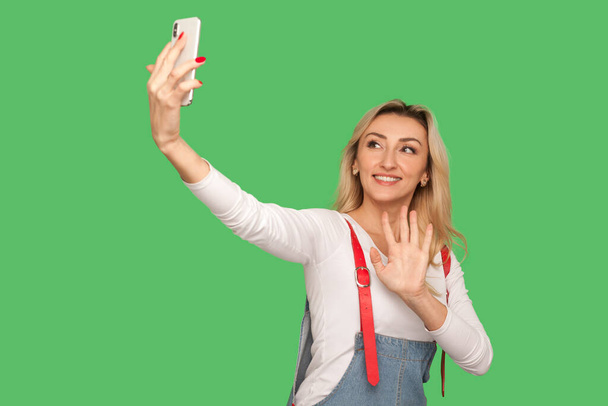Portrait of positive adult woman with friendly smile chatting on video call and waving hello, welcoming subscribers while streaming on smartphone. indoor studio shot isolated on green background - Photo, Image