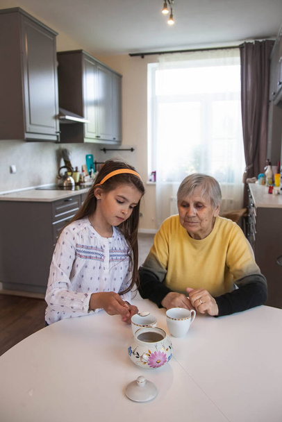 Η γιαγιά και η εγγονή πίνουν τσάι καθισμένοι στο λευκό τραπέζι της κουζίνας. - Φωτογραφία, εικόνα