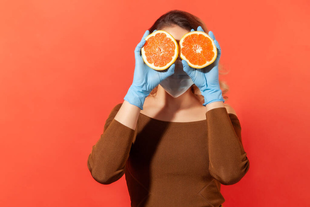 Woman in protective gloves and face mask covering eyes with orange fresh fruits, natural vitamins for immune system and flu treatment, healthy nutrition during quarantine. studio shot, isolated - Foto, Imagem