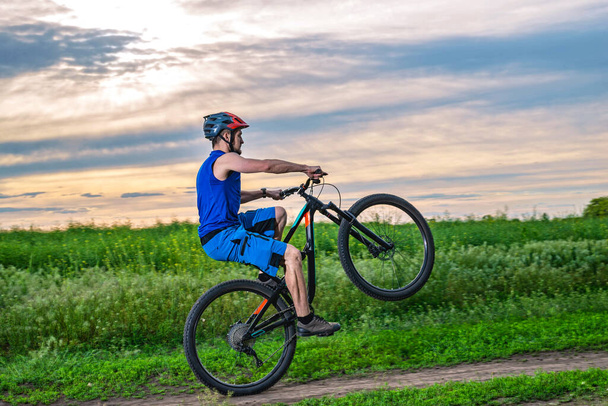 Een fietser met een helm rijdt tijdens zonsondergang op een fiets op het achterwiel. Wielie. - Foto, afbeelding