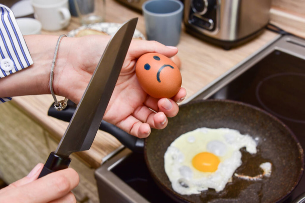 woman in the kitchen in front of the pan holds a knife and breaks a sad smiling egg. - Zdjęcie, obraz