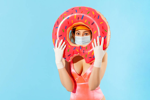 Woman in swimsuit looking through rubber ring, wearing hygienic face mask and surgical gloves to prevent contagious coronavirus on resort beach, rest and summer vacation with covid-19. studio shot - Zdjęcie, obraz