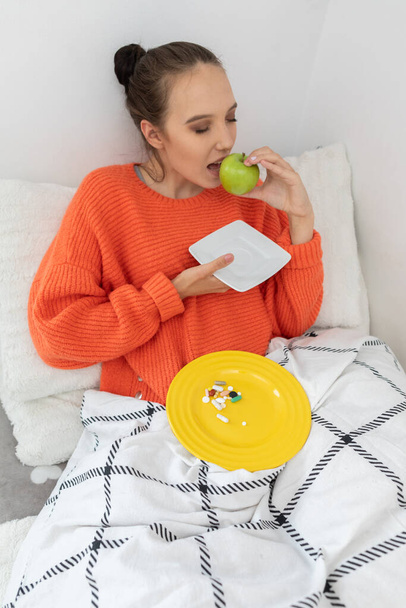 A teenager sits on her bed under the covers in an orange sweater. He's eating a green apple. - Fotó, kép