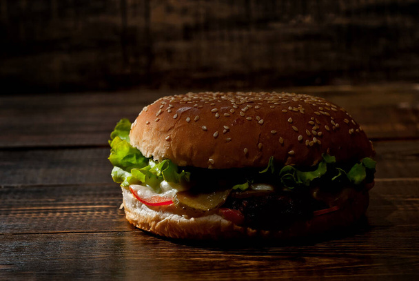 Burger on a dark wooden board. Cheeseburger with cutlet and herbs on black background. Food with contrasting dramatic light. Bun closeup. - Photo, image