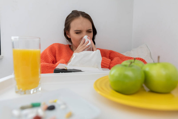 La chica moquea su nariz en un pañuelo. Se sienta enferma en la cama. Hay vitaminas y tabletas y manzanas verdes en el plato
. - Foto, imagen