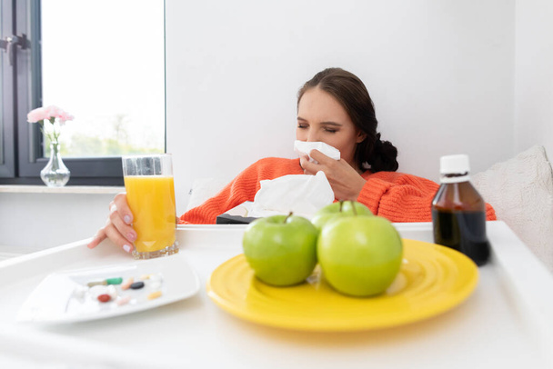 Girl snot her nose in a handkerchief. She sits sick in bed. There are vitamins and tablets and green apples on the plate. - Photo, image
