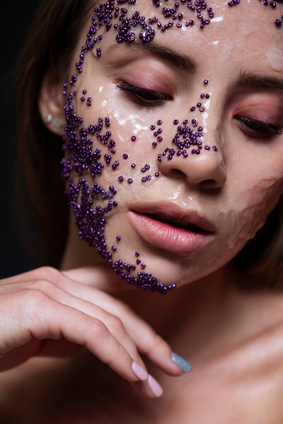 Kreatywna koncepcja piękna. Zbliżenie twarzy kobiety pokryte gliceryną dla efektu mokrego i fioletowe koraliki. Czysta, nieskazitelna skóra. Studio portret na ciemnym tle. - Zdjęcie, obraz