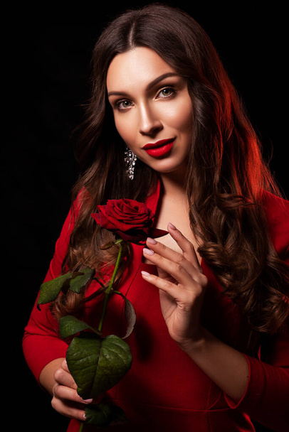 Mulher morena sexy fica em vestido elegante vermelho em um fundo preto. Ela segurando flor de rosa vermelha em suas mãos e sorrir sedutoramente. Conceito de bacharel
. - Foto, Imagem