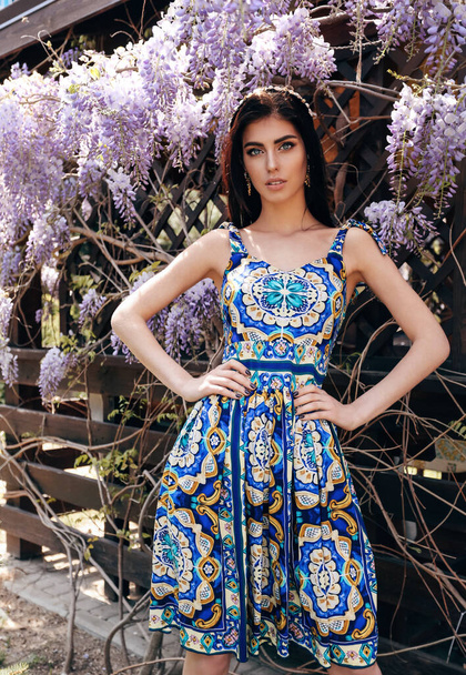 модна зовнішня фотографія красивої чуттєвої жінки з темним волоссям в розкішній сукні з аксесуарами, що позують в літньому саду з квітучими деревами
 - Фото, зображення