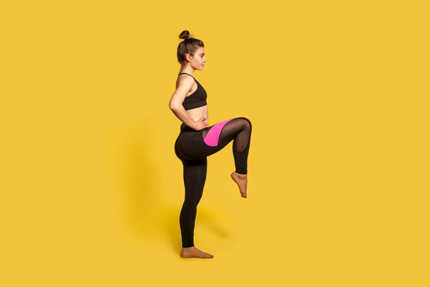 Side view, athletic slim woman with hair bun in tight sportswear doing sport standing with one leg lift, thighs workout, warming up training muscles. full length studio shot, isolated on yellow - Foto, immagini