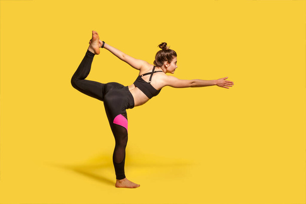Dancer pose. Slim woman with hair bun in tight sportswear practicing yoga, doing Natarajasana exercise on one leg, stretching muscles and balancing. full length studio shot, sport workouts isolated - Фото, изображение