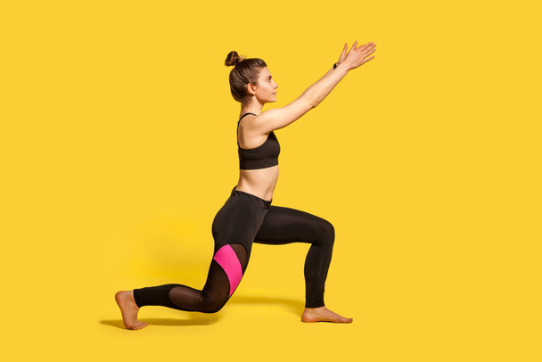Side view, slim young woman with hair bun in tight sportswear doing sport lunge exercise, standing one knee and raising hands, warming up training muscles. full length studio shot, isolated on yellow - Foto, Bild