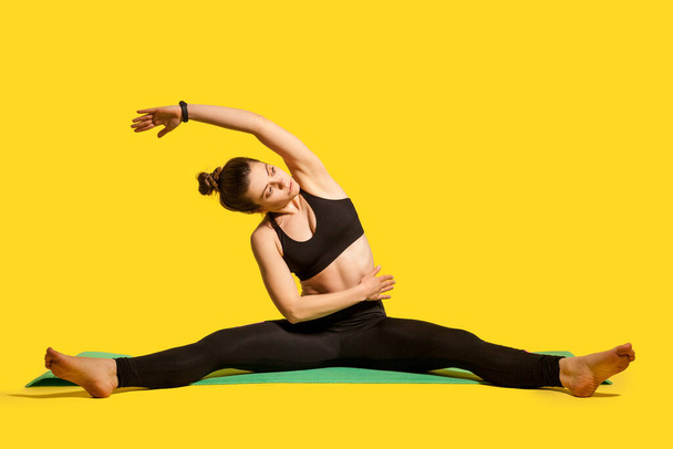 Gymnast girl with hair bun in tight sportswear sitting on mat with spread legs, raising hand bending to side, doing stretching muscle workouts, flexibility exercises. studio shot, isolated on yellow - Photo, image