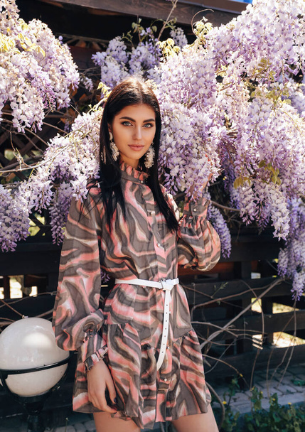 fashion outdoor photo of beautiful sensual woman with dark hair in luxurious dress with accessories posing in summer garden with blooming trees - 写真・画像