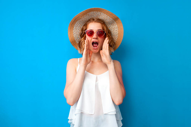summer concept. young crazy girl in a sun hat and glasses shouts on a blue isolated background, a woman speaks loudly in summer clothes on vacation - Photo, image