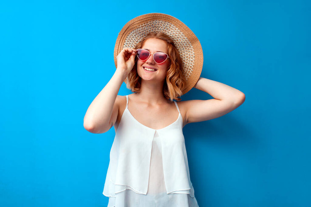 summer concept. portrait of a young girl in a sun hat and glasses on a blue isolated background, a woman in summer clothes on vacation - Photo, Image