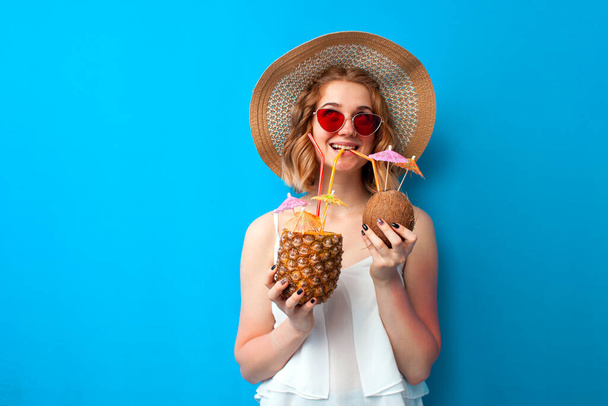 Güneş şapkalı ve gözlüklü genç kız ananas ve hindistan cevizinden yapılmış tropikal kokteyller içiyor ve mavi izole bir arka planda hayaller kuruyor. - Fotoğraf, Görsel