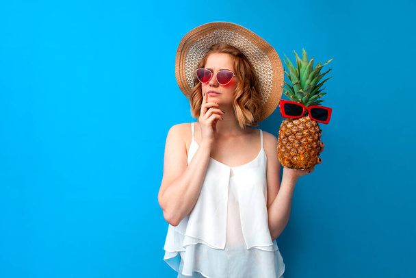 young thoughtful girl in summer clothes with pineapple thinks and dreams about vacation at the resort, woman tourist in summer on a blue isolated background - Foto, Imagen