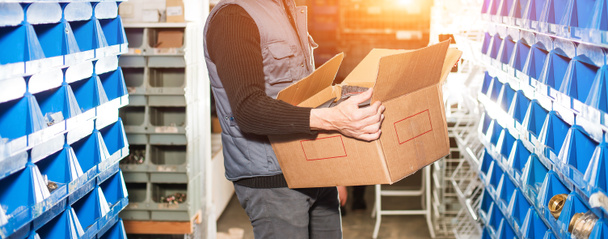 El repartidor en uniforme gris sostiene una caja de pedidos llena en un almacén. Retroiluminación dorada, imagen de pantalla ancha
. - Foto, imagen