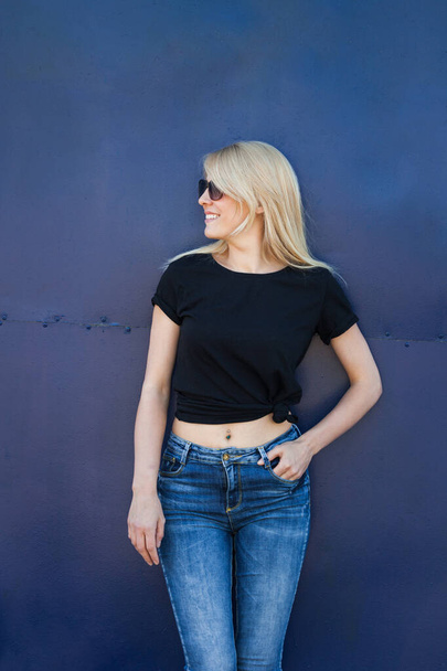 joven rubia sonriente mujer en casual negro camiseta y gafas de sol contra el fondo azul de metal al aire libre en la ciudad camiseta maqueta
 - Foto, imagen