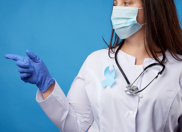 női orvos egy eldobható steril maszk és egy fehér kabát áll a kék háttér, és rámutat a mutatóujj oldalán, egy kék selyem szalag alakú hurkot tűzött a mellkas - Fotó, kép