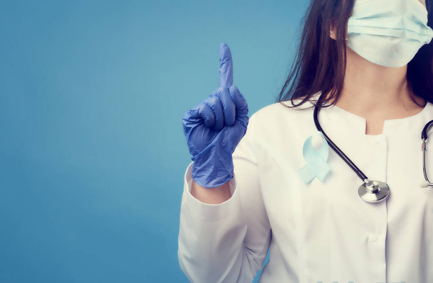 női orvos eldobható steril maszkban, fehér kabátban, kék alapon áll, mutatóujjával felfelé mutat, egy kék selyemszalag van a mellkasára tűzve. - Fotó, kép