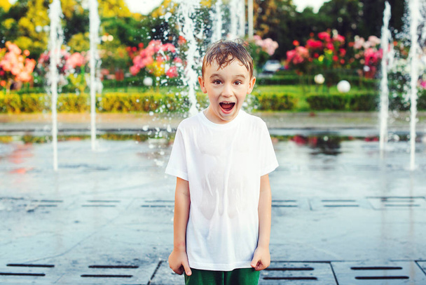 Ein fröhlicher Junge spielt an einem sonnigen Sommertag auf dem Platz zwischen den Wasserstrahlen im Brunnen. Sommerurlaub, Lifestyle. Junge wird mit Wasser aus dem Brunnen gekühlt. - Foto, Bild