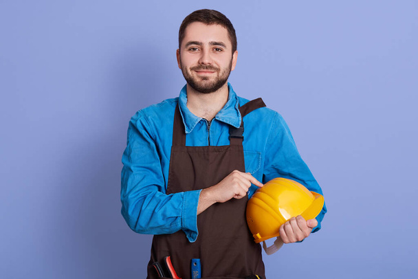 Portrait of delighted hard working glad young man looking directly at camera, holding yellow helmet in one hand, wearing blue overall and brown apron, posing isolated over blue background in studio. - Photo, Image