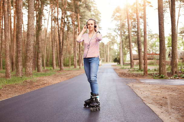 Portrait of confident sporty energetic young female listening to music, putting on headphones, having roller blades on legs, enjoying weekends, having rest, being fond of nature. Lifestyle concept. - Zdjęcie, obraz