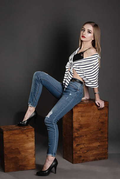 a girl in a striped sweater and blue city jeans on a wooden box on a gray background, a full-length photograph, the girl put her foot on the box, - Foto, Bild