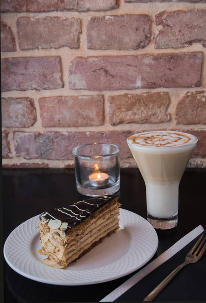 Ένα κομμάτι κέικ σε ένα λευκό πιάτο - με ένα latte σε ένα ψηλό ποτήρι σε ένα μαύρο τραπέζι με ένα αναμμένο κερί, στο φόντο ενός κόκκινου τοίχου από τούβλα. - Φωτογραφία, εικόνα