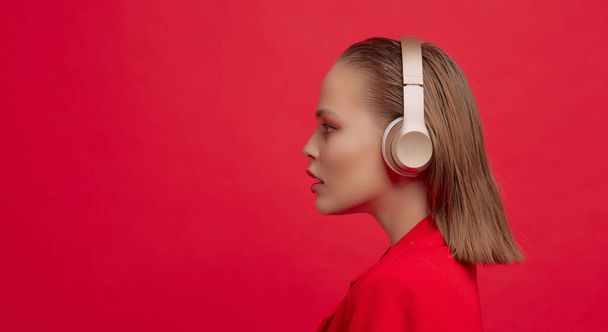 A cute young girl with a beautiful hairstyle is listening to music in wireless headphones on a red background. - Fotó, kép
