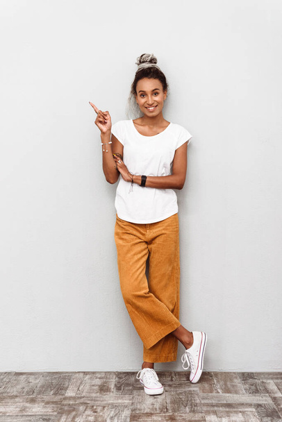 Full length of a smiling young african woman wearing casual outfit standing isolated over white background, pointing finger away at copy space - Φωτογραφία, εικόνα
