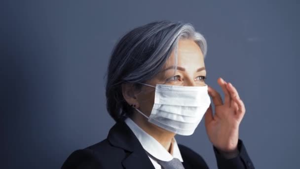 White-haired business woman adjusts a protective mask while standing in the office against the background of a dark wall. Business during the pandemic quarantine period. Business concept. Prores 422 - 映像、動画