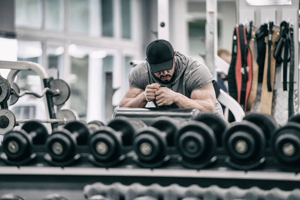 heavy stress training concept of young bearded tired male in black cap resting after hard workout in fitness indoor gym among iron dumbbells - 写真・画像
