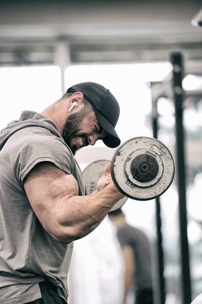 sport motivation concept of strong young athlete man with beard lifting weight barbell pumping iron training biceps with angry emotions pushing to limits - Foto, afbeelding