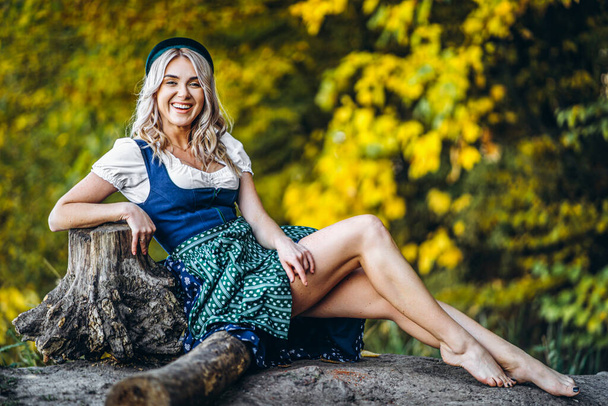 Oktoberfest, St. Patrick day, intenational beer day. Barefoot happy pretty blond girl in dirndl, traditional beer festival dress, sitting outdoors with blured colorful trees behind - Fotografie, Obrázek