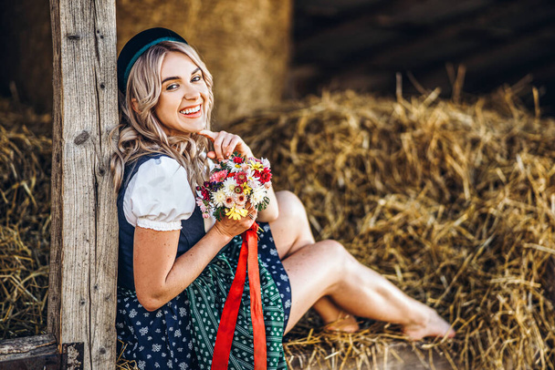 Cute blonde in dirndl, traditional festival dress with bouquet of field flowers sitting on the wooden fence at the farm with haystacks behind - Fotografie, Obrázek