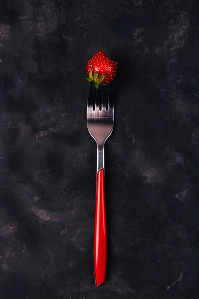One strawberry on a red fork in the center of the black rustic background. Top view. Vertical. Diet minimal concept. - Foto, Bild