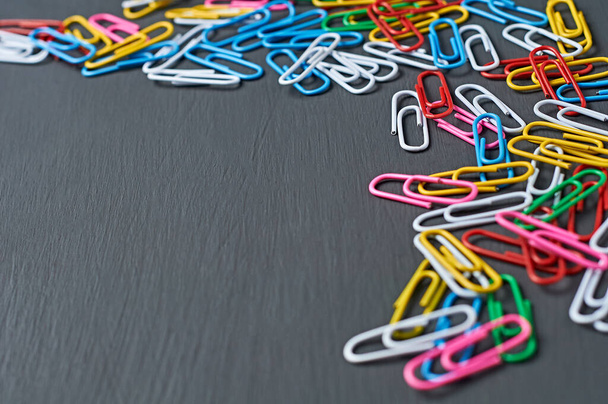 Lot of scattered different colors paper clips red, green, blue, yellow, white and pink for office work or education lies in corner on dark scratched concrete table. Space for text - Foto, Bild