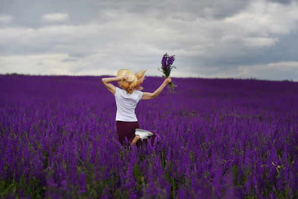 Una ragazza in un cappello di paglia con un mazzo di fiori selvatici corre lungo un lussureggiante campo di lavanda fiorita con fiori
. - Foto, immagini