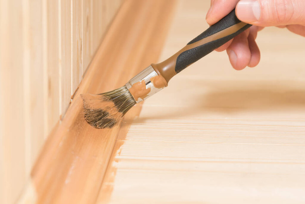 a small brush covers the skirting board of the wooden floor with dark varnish - Zdjęcie, obraz