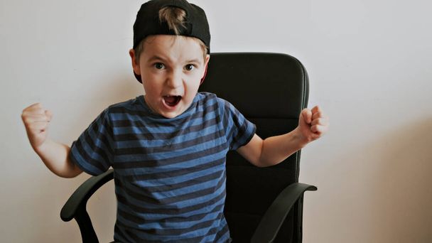Хлопчик сидить у чорному офісному кріслі і кричить як переможець
 - Фото, зображення