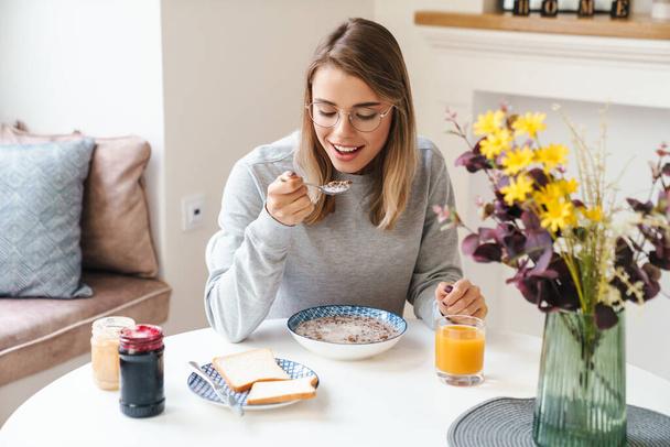 Photo of cheerful young woman in eyeglasses eating cereal while having breakfast at living room - Foto, afbeelding